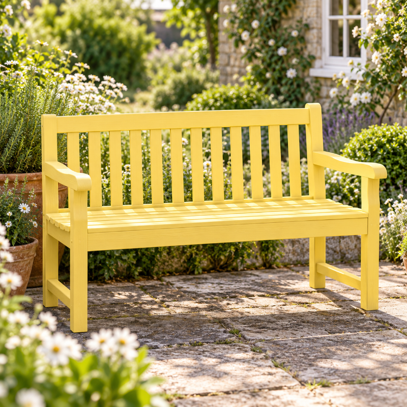 A bright yellow wooden garden bench with armrests and vertical slats, placed on a stone patio in a lush garden setting with flowers and greenery.
